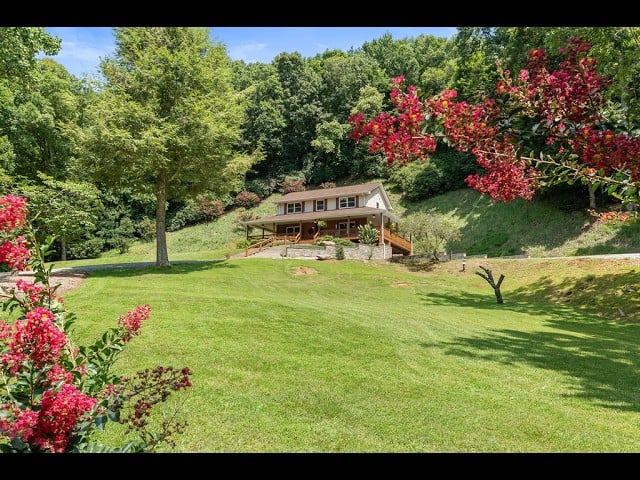 House on a hillside above a broad lawn, framed by mature trees and pink flowering shrubs.