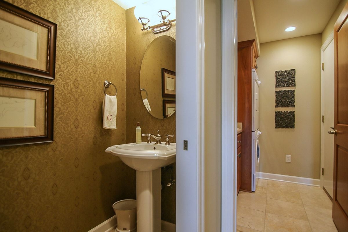 Bathroom with gold-patterned wallpaper, a pedestal sink and an adjoining tiled laundry hallway.