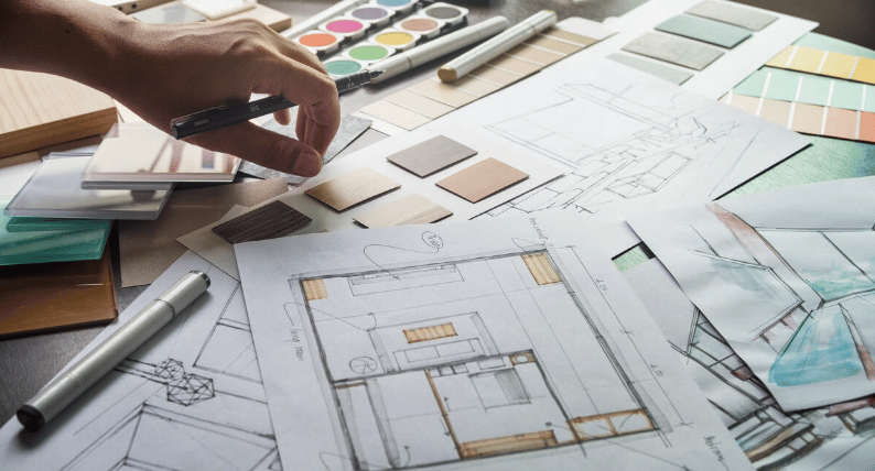 A hand selects a finish sample among architectural plans, paint swatches, and material samples.