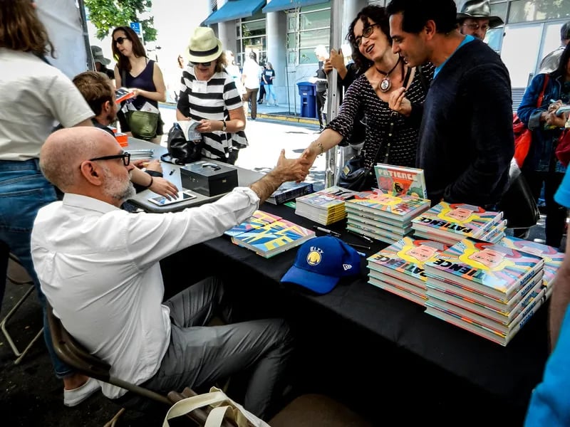 A lively outdoor book signing at the Bay Area Book Festival. Acclaimed cartoonist Daniel Clowes, a bald man with glasses and a white button-down shirt, sits at a black table piled with copies of his graphic novel 'Patience.'