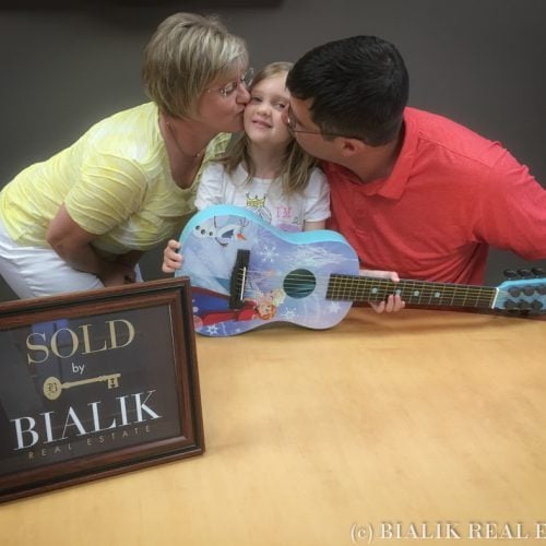 Parents kiss their daughter as she holds a toy guitar beside a SOLD sign.