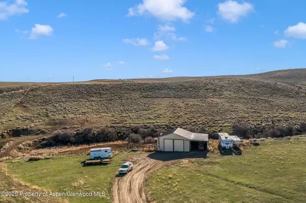 Aerial view of a rural property with a metal outbuilding, trailers, dirt drive, and dry hillside.