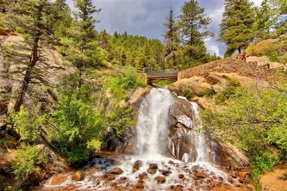A waterfall tumbles over rocky ledges beneath a footbridge in a forested mountain landscape.