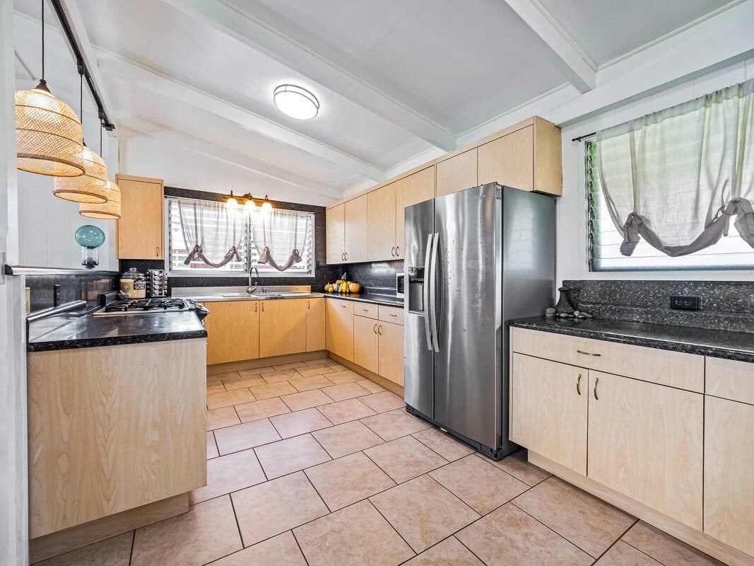 Kitchen with light wood cabinets, dark countertops, stainless steel refrigerator, and large tile floor.