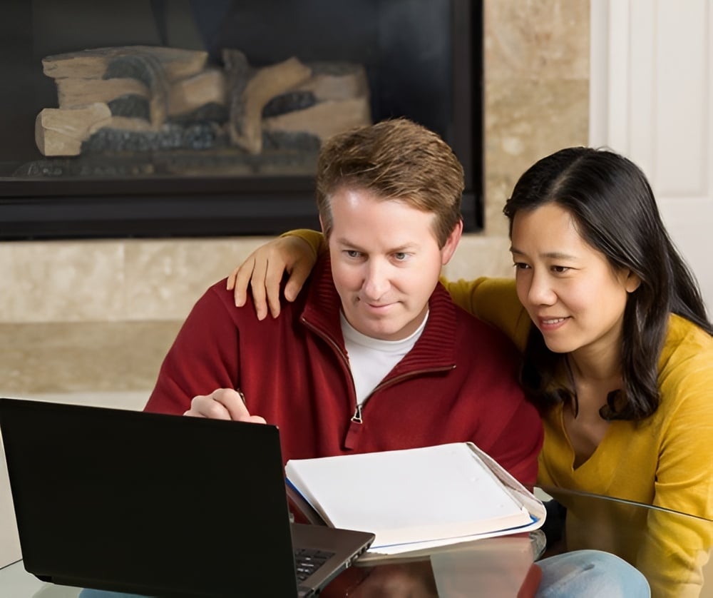 A couple looks at a laptop together, with an open binder on the table.