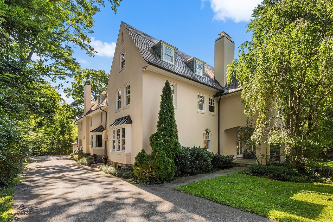 Cream-colored house with slate roofs and dormers beside a tree-lined driveway and lawn.