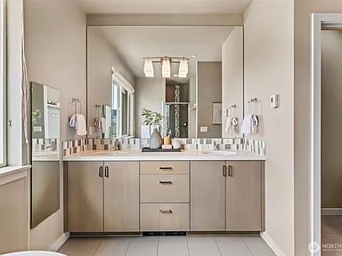 Bathroom with a long double vanity, broad mirror, tiled backsplash, and windows along the left wall.