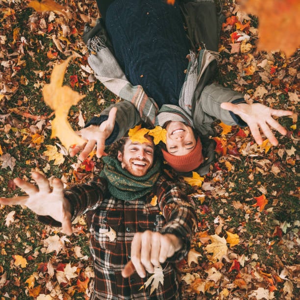 Two smiling people lie in colorful autumn leaves and toss leaves toward the camera.