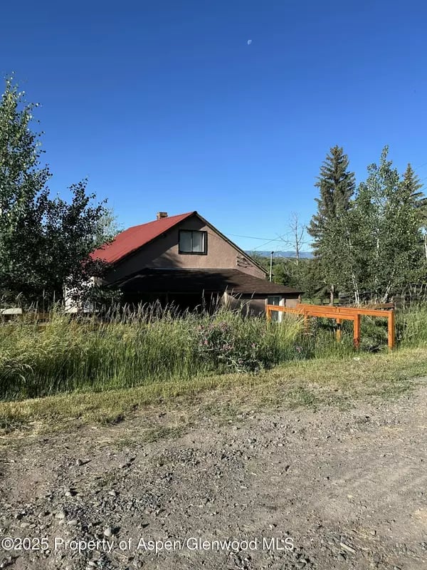 Brown house with a red metal roof, partly screened by tall grasses and trees beside a gravel road.