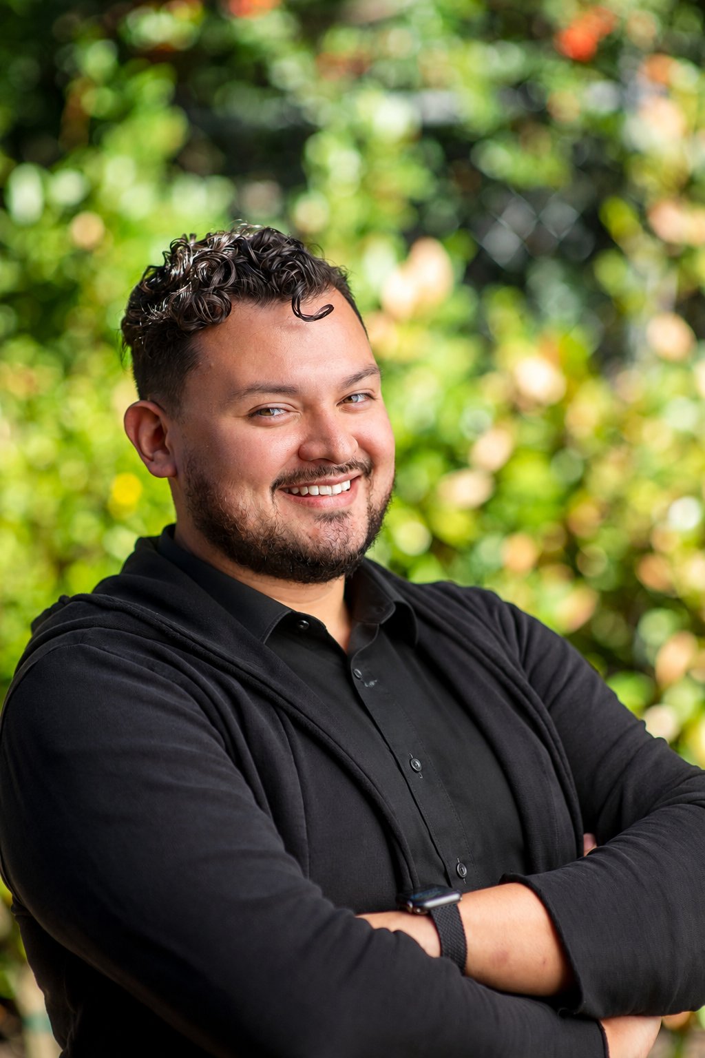 Wilbur Canizalez smiling with his arms crossed in front of blurred greenery.