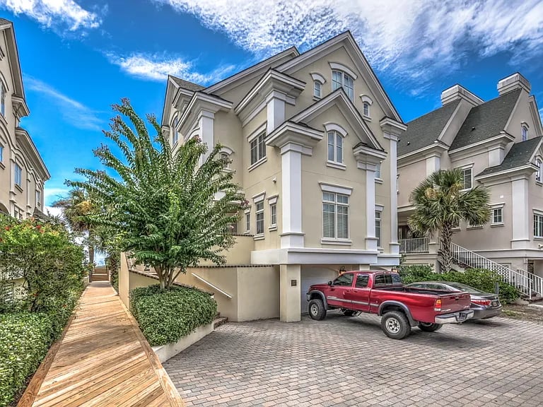 Multi-story beige home with white trim, a red pickup, and a wooden walkway beside it.