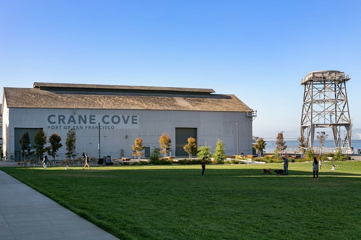 Crane Cove Park in San Francisco’s Dogpatch neighborhood, featuring a grassy waterfront lawn, the historic Crane Cove warehouse, and former shipyard crane overlooking the Bay.