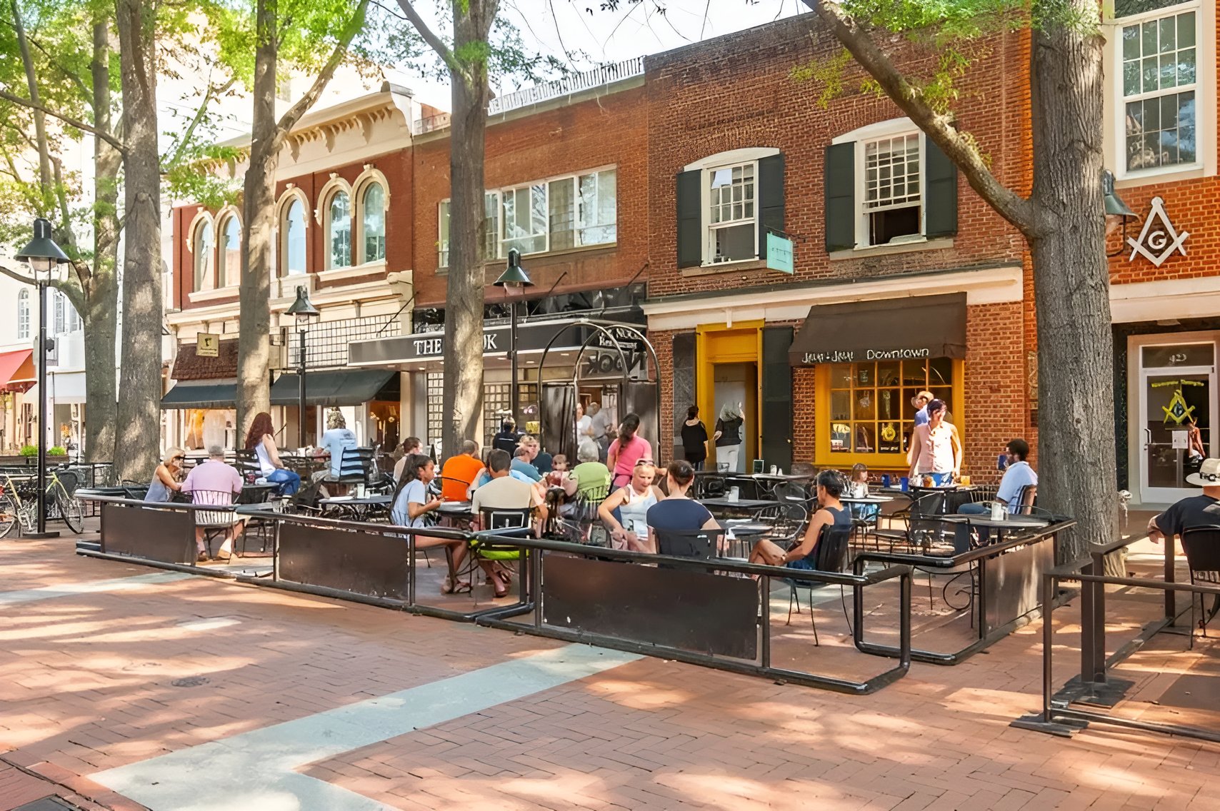 Downtown Charlottesville storefronts and a busy outdoor dining area shaded by mature trees.