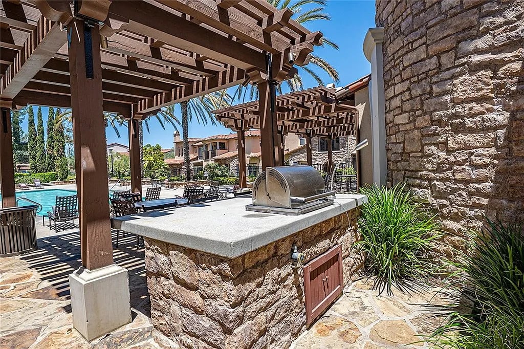 Poolside barbecue grill beneath wooden pergolas, with stone counter and palm trees.