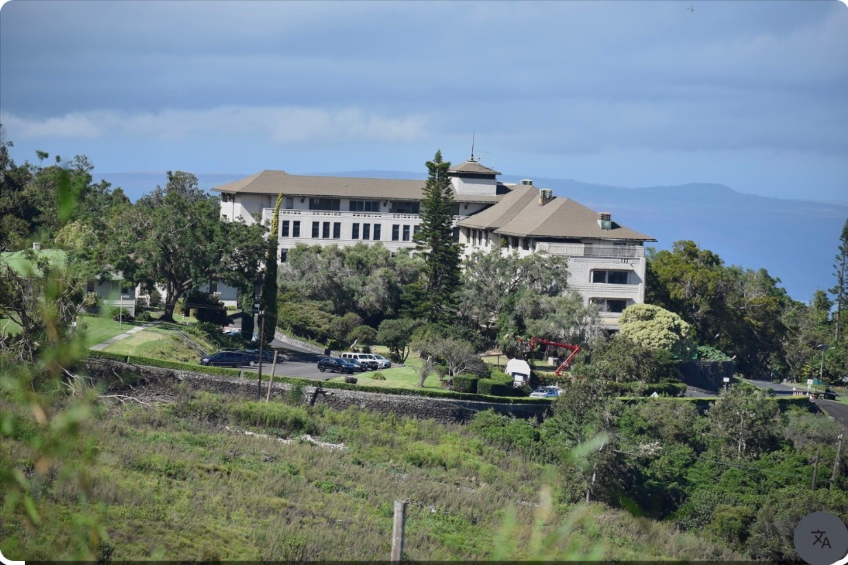 Large hillside building surrounded by trees and landscaped grounds, with ocean and distant ridges beyond.