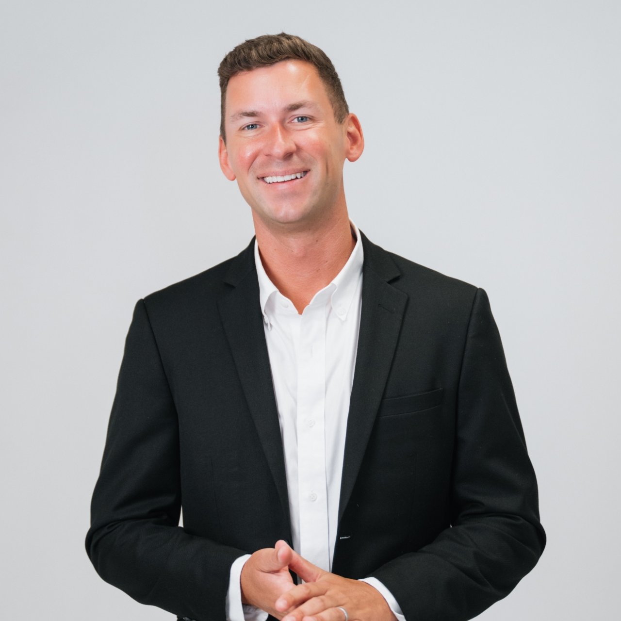 Smiling man in a black suit jacket and white shirt, hands clasped.