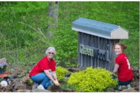 Two women in red shirts beside shrubs and flowers near a wooden Habitat sign.