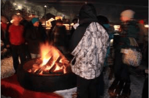 People gather around a large fire pit outdoors at night.
