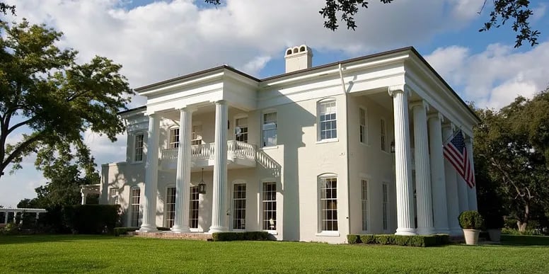 White Greek Revival mansion with tall columns, a balcony, and an American flag.