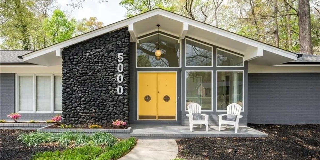 Mid-century modern home with yellow double doors, a dark stone facade, and two white chairs at the entrance.