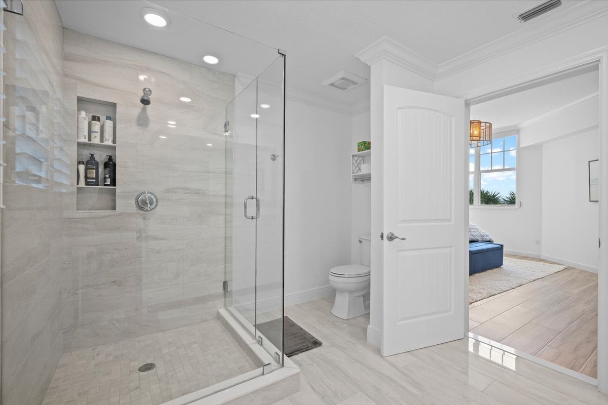 Condominium bathroom with a glass-enclosed walk-in shower and pale tile, opening onto a bedroom.