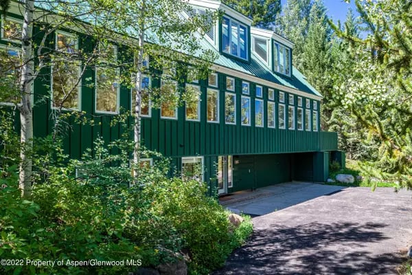 Multi-level dark green home with many windows and a driveway amid mature trees.