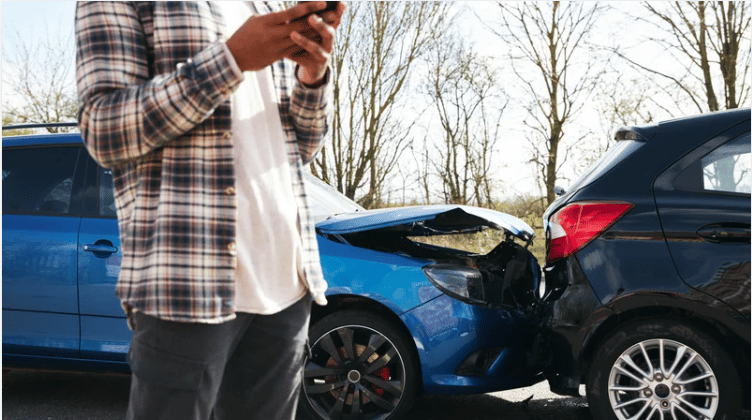A person checks a phone beside a damaged blue car and a black SUV.