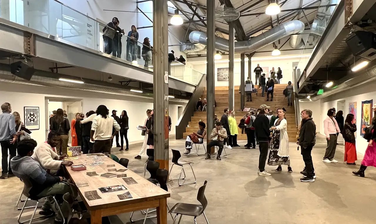 A modern gallery with a high ceiling, filled with people at an art event. A wooden table with papers, chairs, and a staircase to a second level are visible, with various artworks on the walls.