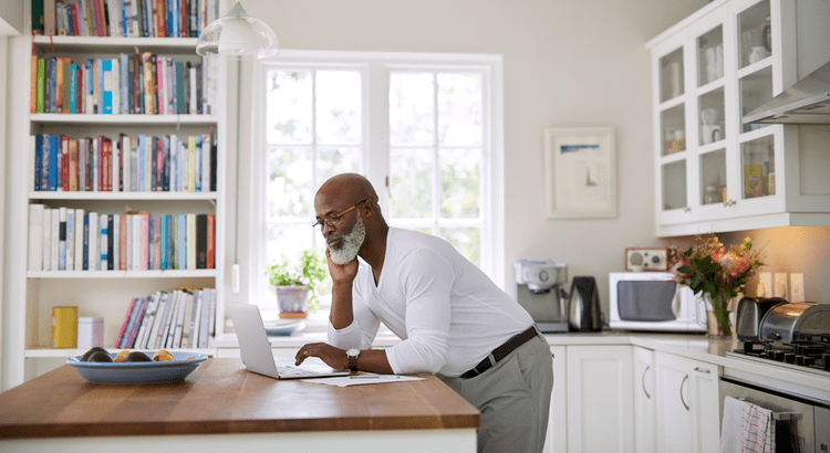 A man in a white shirt is working on a laptop while seated at a kitchen table.