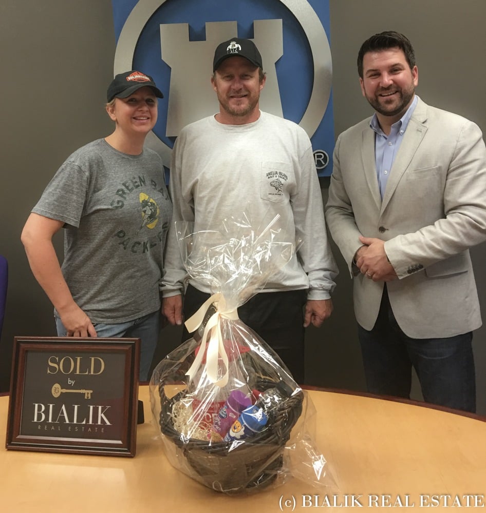 Three adults pose with a gift basket beside a SOLD by Bialik Real Estate sign.
