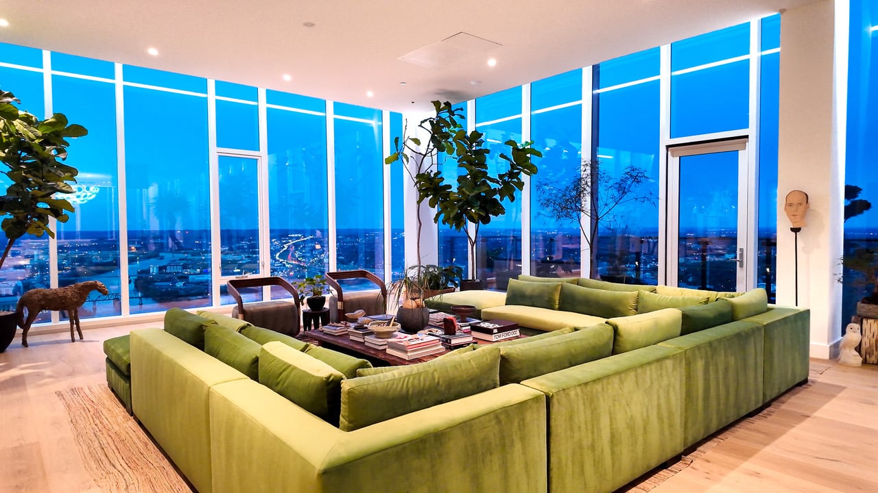 High-rise living room with a large green sectional and floor-to-ceiling windows overlooking the city at dusk.