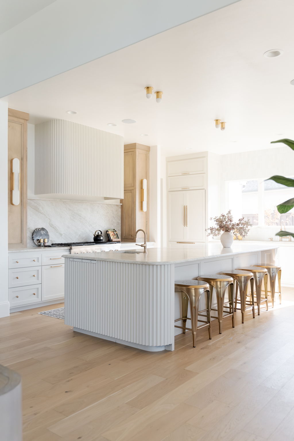 Bright kitchen with a fluted white island, pale wood cabinets, and gold bar stools.