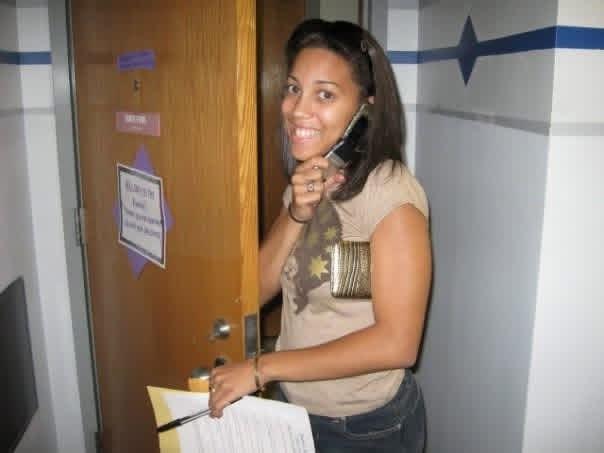 A photo of Arielle with a flip phone and checklist standing in front of a door to her dorm room.