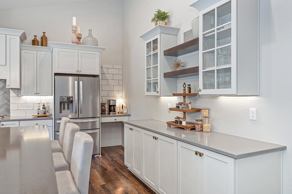 Bright white kitchen with glass-front cabinets, stainless steel refrigerator, and long counter with seating.