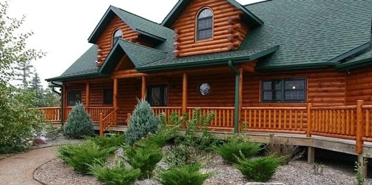 Two-story log home with a covered porch, arched dormer windows, and landscaped gravel beds.