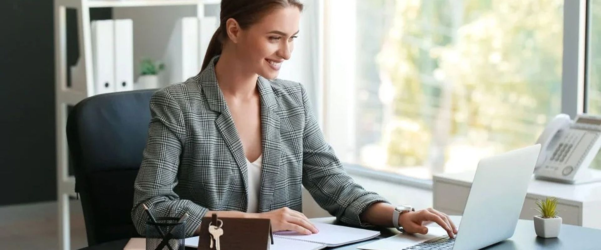 Woman working at an office desk with a laptop, paperwork, and keys.