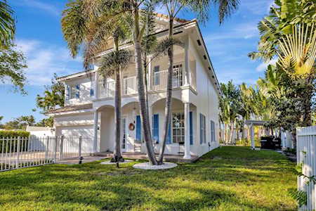 Two-story white house with balconies, palm trees, and a white fence along the lawn.