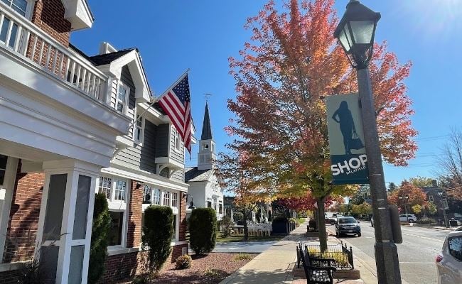 Downtown Delafield street with shops, an American flag, autumn trees, and a church steeple on a sunny day.