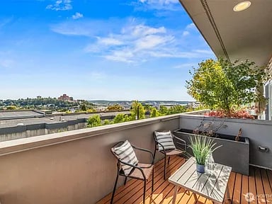 Apartment balcony with two chairs and a potted plant, overlooking rooftops and distant water.
