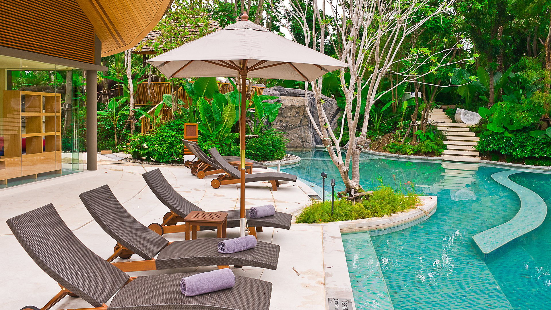 Outdoor pool patio with lounge chairs and umbrella beside a modern glass-walled home.