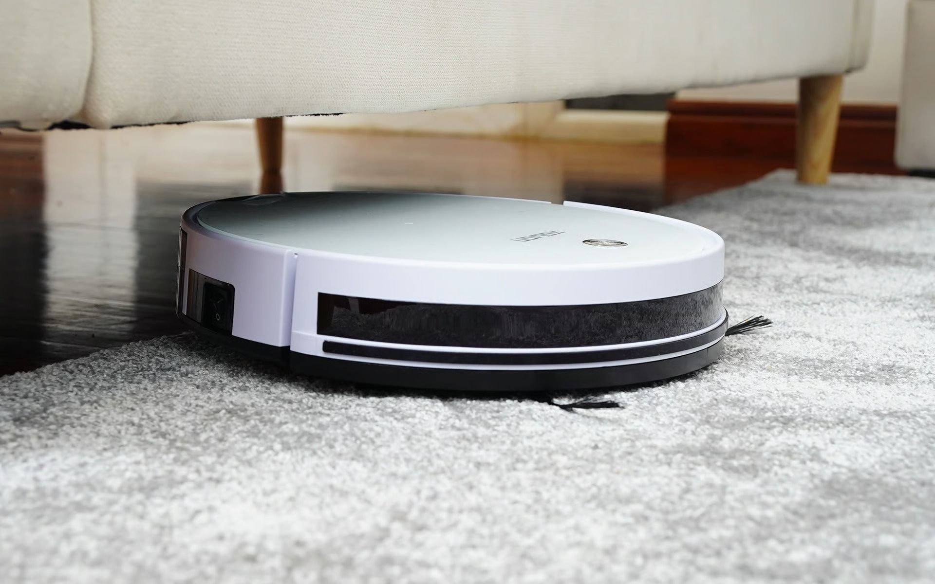White robot vacuum cleaner on a gray carpet beneath a sofa.