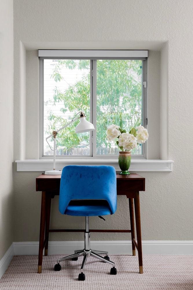Home office nook with a blue swivel chair, wood desk, and leafy window view.
