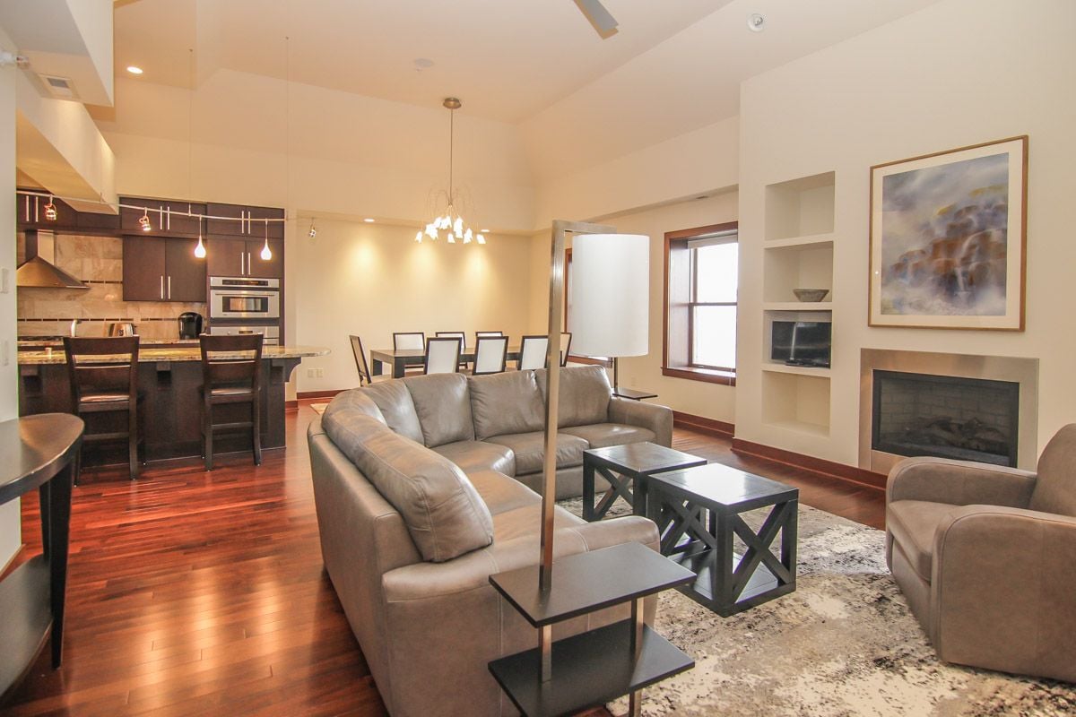 Open-plan condo living room with a gray sectional, fireplace, built-in shelves, and adjoining kitchen.