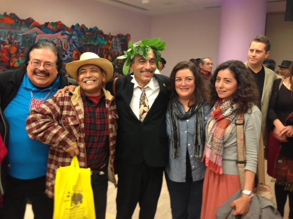 San Francisco Poet Laureate Alejandro Murguía, wearing a dark suit and a leaf crown, poses with a group of four other smiling adults, including two men in casual attire and two women in scarves, at an indoor event with an abstract, colorful mural in the background.
