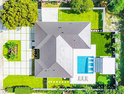 Aerial view of a home with a blue pool, paved patio, and mature trees.