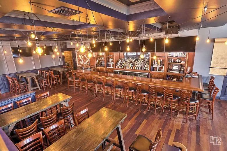 Restaurant bar with a long wooden counter, pendant lights, and rows of wooden tables and chairs.