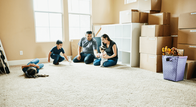A family of four sits in a sunlit room filled with moving boxes. Parents sit on the carpet with a baby, while a young boy plays nearby. The scene feels warm and cheerful.A family of four sits in a sunlit room filled with moving boxes. Parents sit on the carpet with a baby, while a young boy plays nearby. The scene feels warm and cheerful.