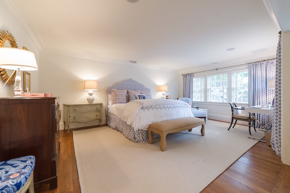 Master bedroom with a patterned bed, tan bench, hardwood floors, and a desk beside large windows.