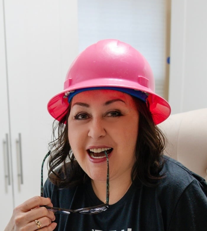 Woman in a pink hard hat holding eyeglasses near her mouth.