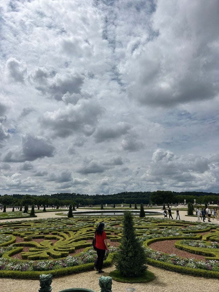 Formal garden with clipped parterre hedges, flower beds, a reflecting pool, and visitors beneath a cloudy sky.